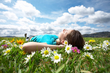 A woman lies peacefully in a field of flowers, her eyes closed, under a blue sky with fluffy white clouds.