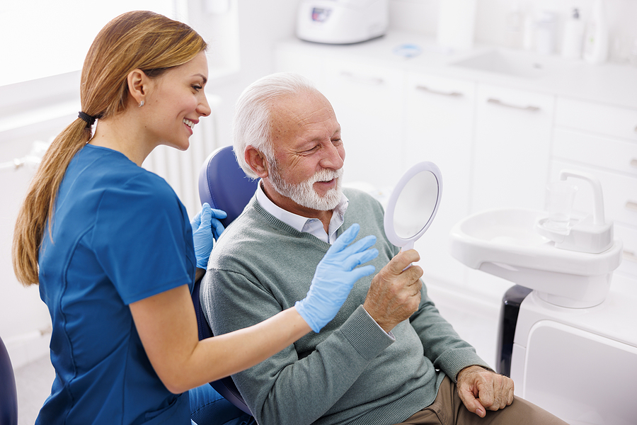 A senior man in a dental chair smiles as he looks at himself in the mirror with a dental professional assisting beside him.